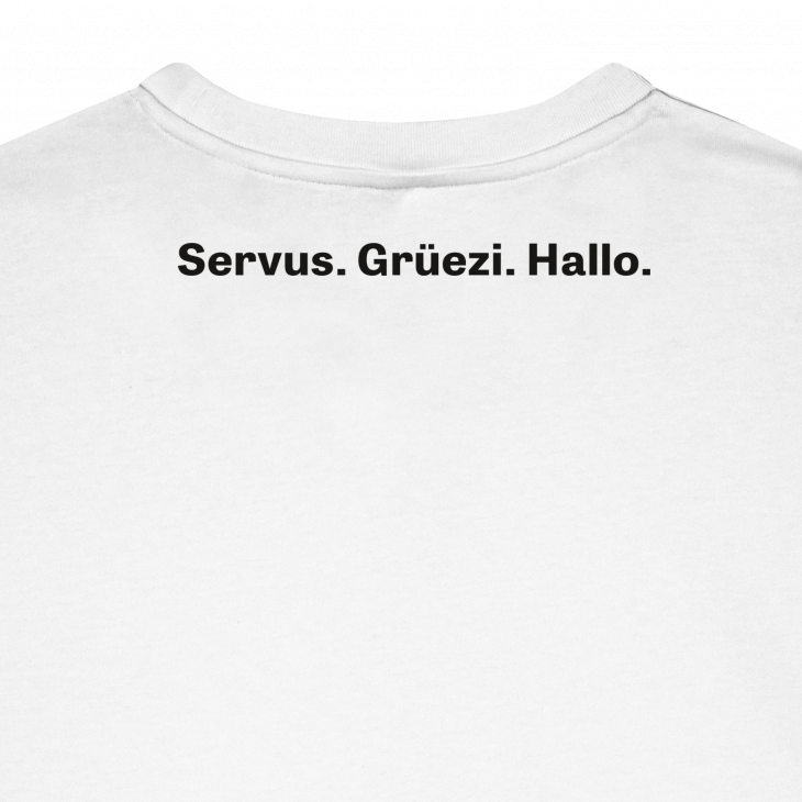 Das Bild zeigt die Rückseite eines weißen T-Shirts mit dem schwarzen Schriftzug 'Servus. Grüezi. Hallo.' am oberen Rücken. Das Shirt vermittelt einen freundlichen Gruß aus Deutschland, Österreich und der Schweiz und ist schlicht sowie modern gestaltet.