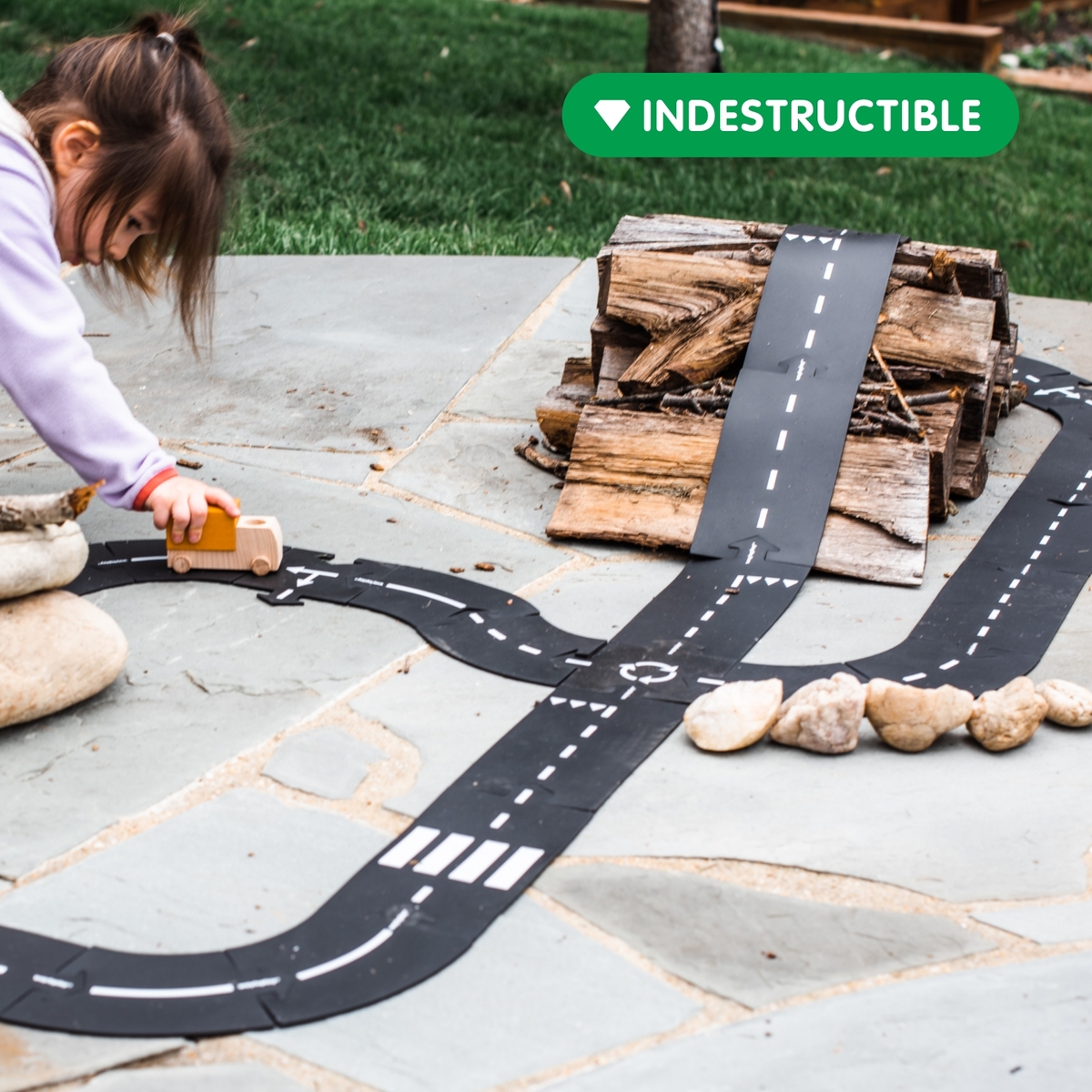 Ein Kind spielt im Freien mit einem flexiblen Straßenbau-Set auf Steinplatten. Die schwarze Spielstraße ist zu einem Straßenverlauf zusammengesetzt und wird mit Steinen und Holz ergänzt. Das Kind schiebt ein Spielzeugauto über die Piste.