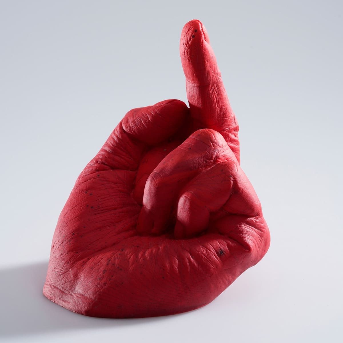 Eine rote, glasartige Plastikskulptur in Form einer Hand mit ausgestrecktem Mittelfinger auf einem weißen Hintergrund. Das Kunstwerk trägt den Titel 'Study of Perspective in Glass' von Ai Weiwei.