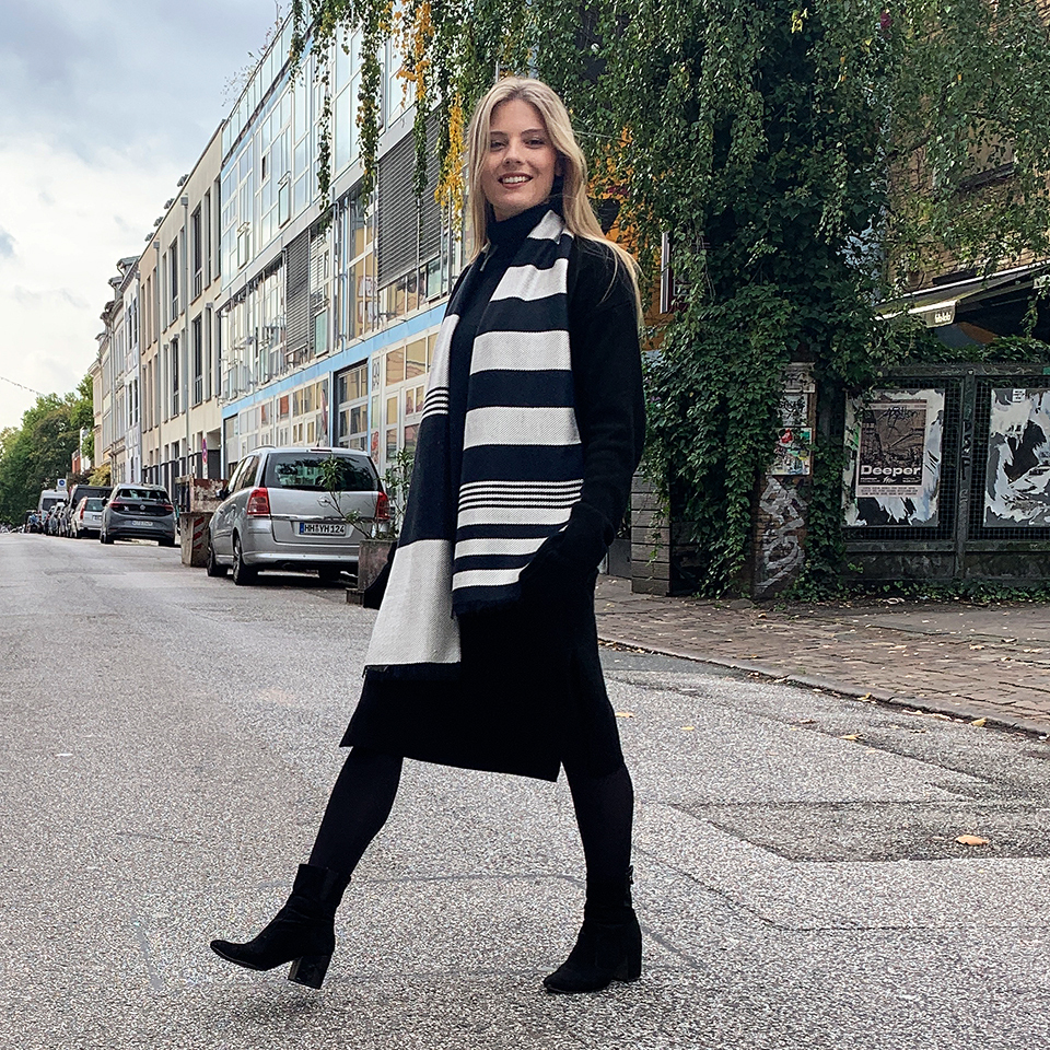 Eine Frau steht auf einer städtischen Straße und trägt einen schwarz-weiß gestreiften Seidenschal. Im Hintergrund sind geparkte Autos und Gebäude zu sehen.