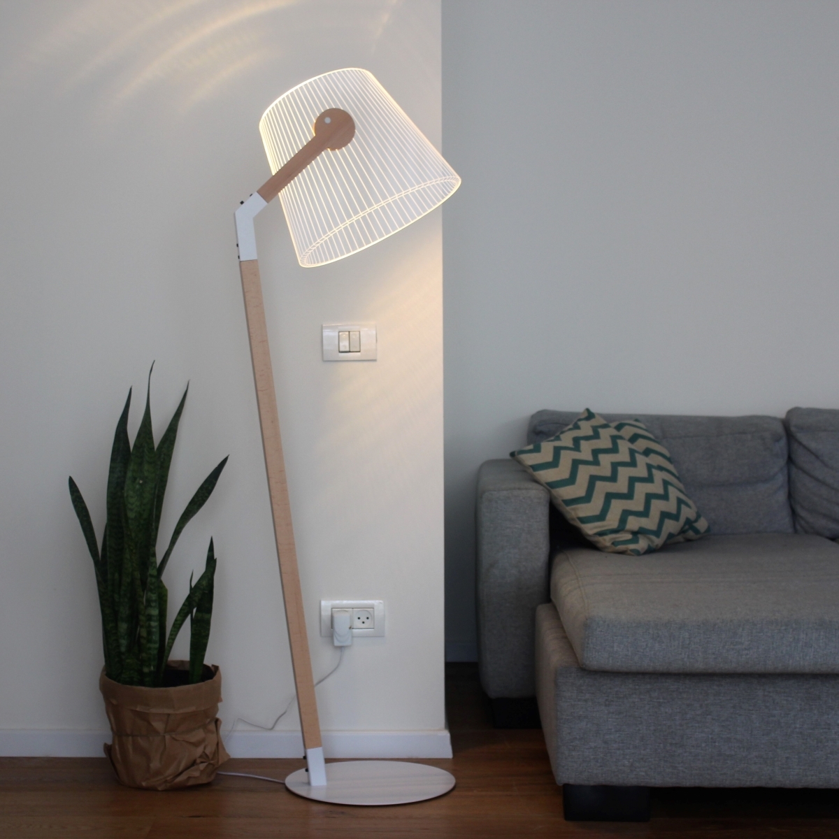 Minimalistische Stehlampe füllt die Ecke eines Wohnzimmers und beleuchtet einen modernen, grauen Sofa-Bereich. Die Lampe hat weiße und holzfarbene Elemente. Neben dem Sofa befindet sich eine grüne Pflanze in einem braunen Topf.