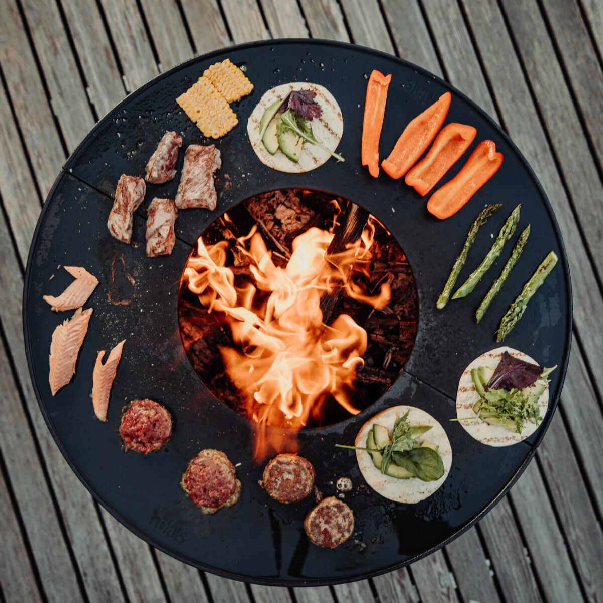 Runde Plancha Halbring Feuerstelle von BOWL mit brennendem Feuer in der Mitte, umgeben von verschiedenen Speisen wie Gemüse, Fleisch und Brot auf einem Holzboden. Ideal für Grillen im Freien und Outdoor-Kochen.