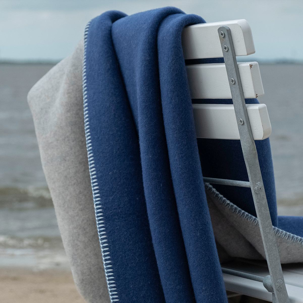 Eine weiche Merino-Decke in dunklem Blau und dezentem Hellgrau liegt über einer weißen Holzlehne eines Stuhls am Strand mit Blick auf das Meer. Die Szene wirkt ruhig und einladend, ideal für entspannte Momente im Freien oder zum Kuscheln im Haus.