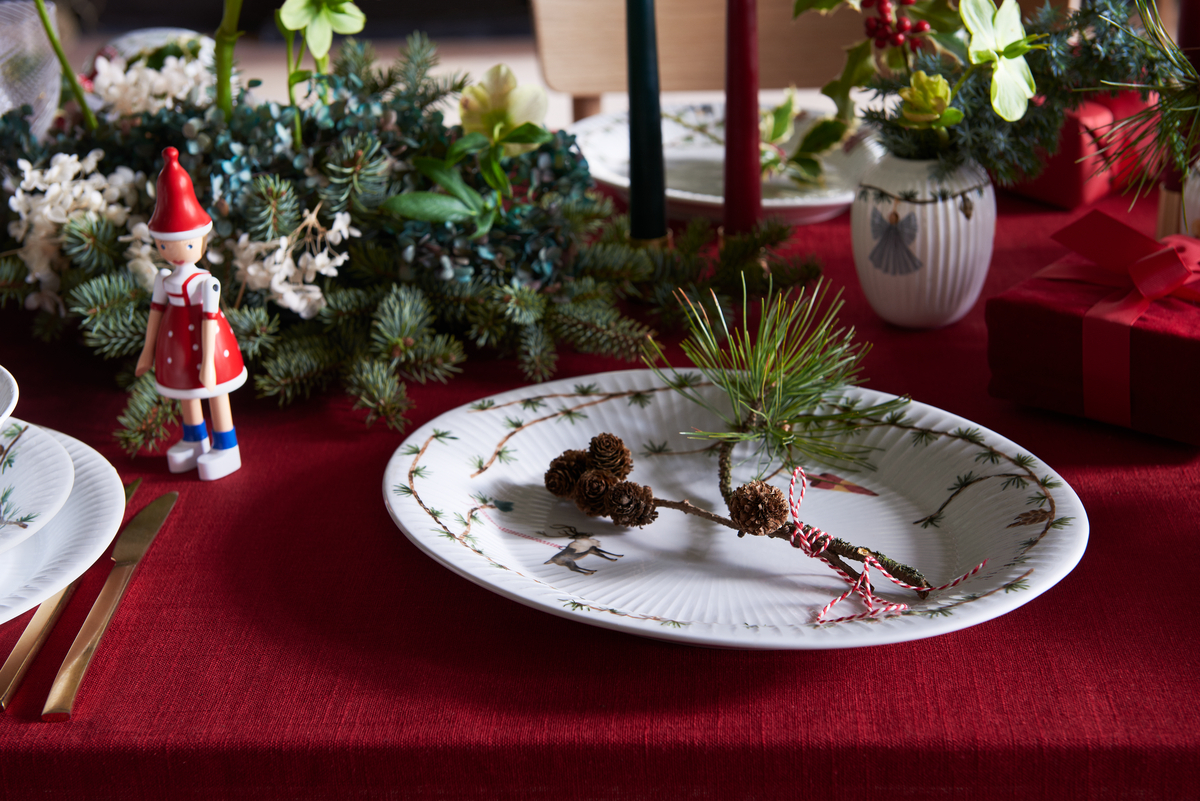 Ein festlich gedeckter Tisch mit einer Weihnachtsplatte »Hammershøi«, dekoriert mit einem Kiefernzweig und Zapfen. Daneben steht eine kleine Weihnachtsmannfigur. Im Hintergrund sind eine Vase mit Blumen, Kerzen und ein rotes Geschenk auf einem roten Tischtuch.