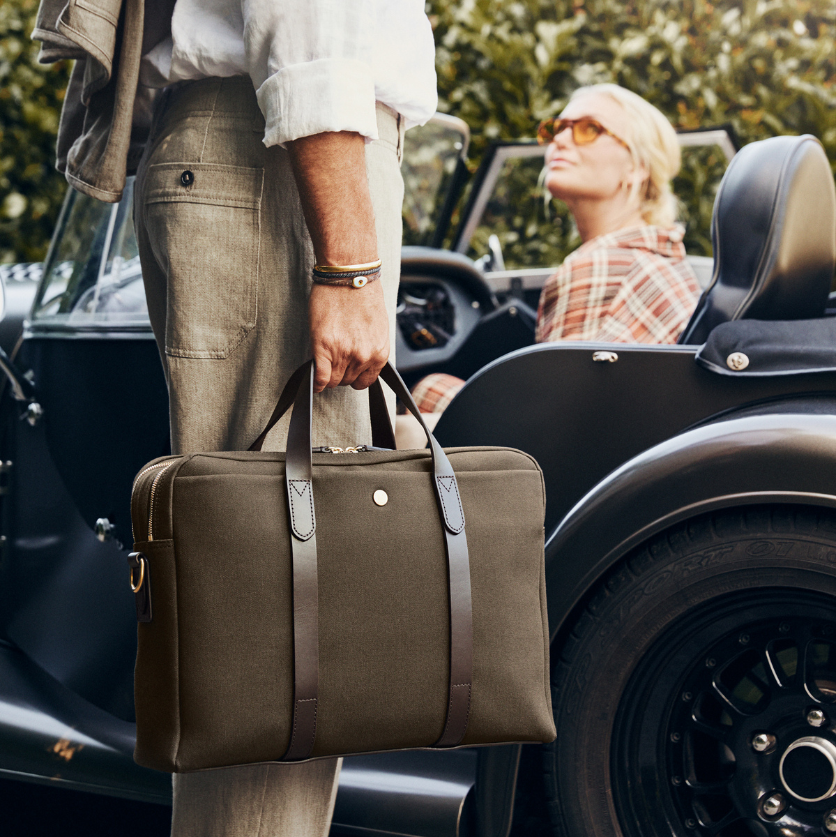 Ein Mann hält eine dunkelgrüne Businesstasche »ENDEAVOUR« von Mismo mit braunen Ledergriffen. Er steht neben einem schwarzen Cabrio. Eine Frau mit Sonnenbrille sitzt im Auto. Im Hintergrund sind grüne Hecken sichtbar.