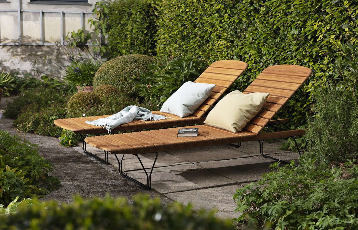 Zwei hölzerne Gartenliegen mit verstellbaren Rückenlehnen stehen auf einer Terrasse, umgeben von sorgfältig gepflegten Hecken und anderen grünen Pflanzen. Eine der Liegen hat ein helles beiges Kissen, die andere ein blasses grünes Kissen. Auf einer Liege liegt eine gefaltete Decke, auf der anderen ein Buch.