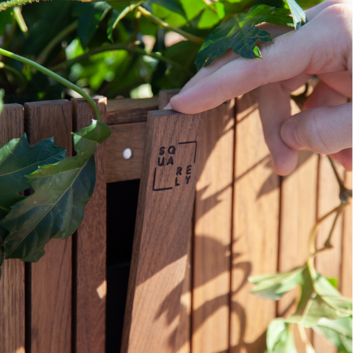 Nahaufnahme eines Pflanzgefäßes »GrowWIDE« in Holzoptik mit grünen Blättern. Eine Hand berührt das Gefäß, auf dem das Logo 'SQUARELY' zu sehen ist.