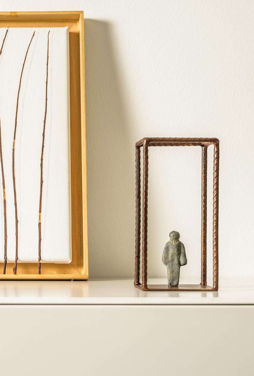 Eine minimalistische Skulptur namens 'The Window Room' von Fantini, Erli. Die Skulptur zeigt eine kleine, abstrahierte menschliche Figur, welche von einem rechteckigen Metallrahmen umgeben ist. Der Hintergrund ist weiß, und neben der Skulptur befindet sich ein gerahmtes Kunstwerk mit vertikalen Linien.