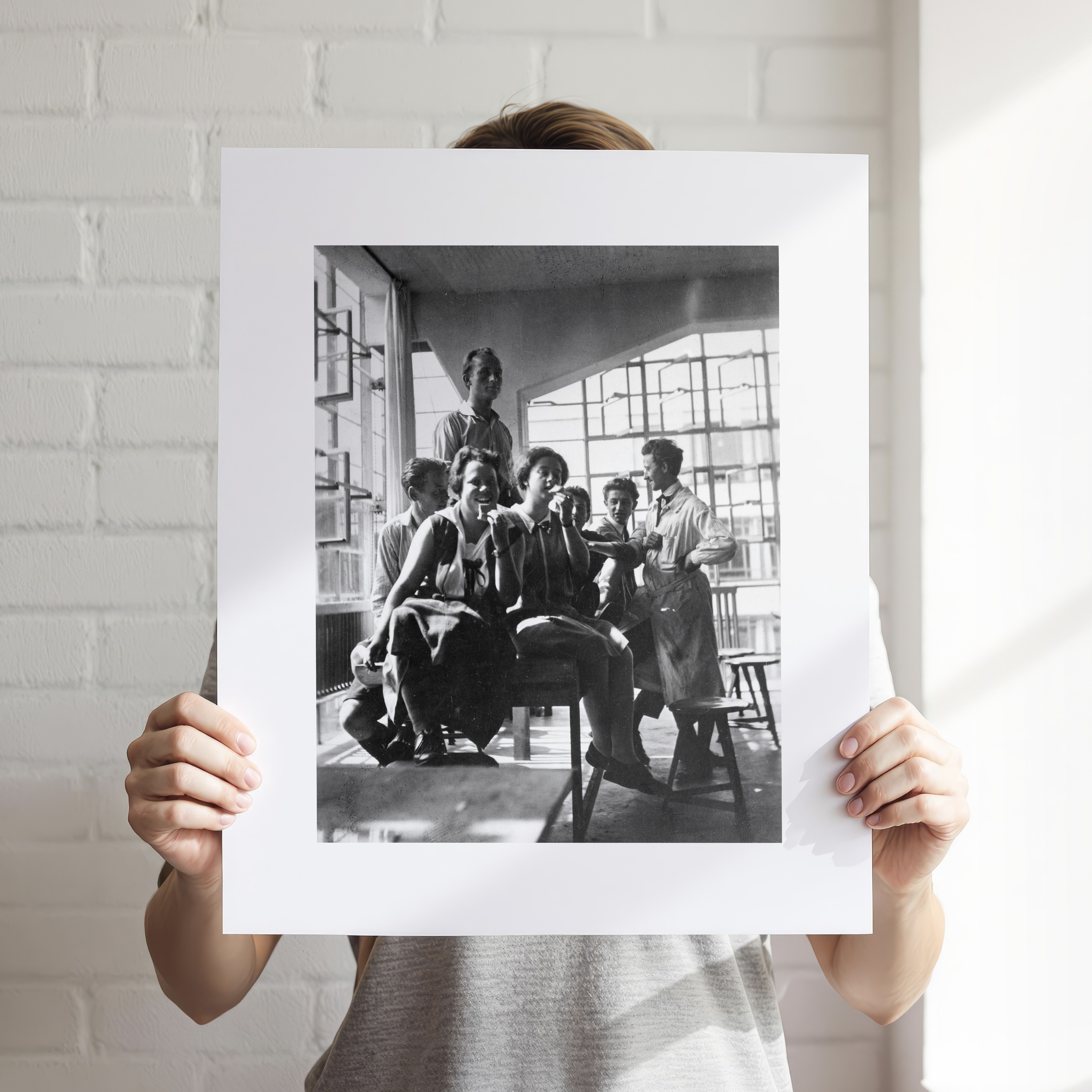 Eine Person hält einen Kunstdruck mit einer Schwarzweißfotografie, die eine Gruppe von Menschen im Bauhaus-Stil zeigt. Im Hintergrund sieht man eine weiße Backsteinwand. Der Druck ist modern gestaltet und erinnert an die künstlerische Ära des Bauhauses in Berlin.