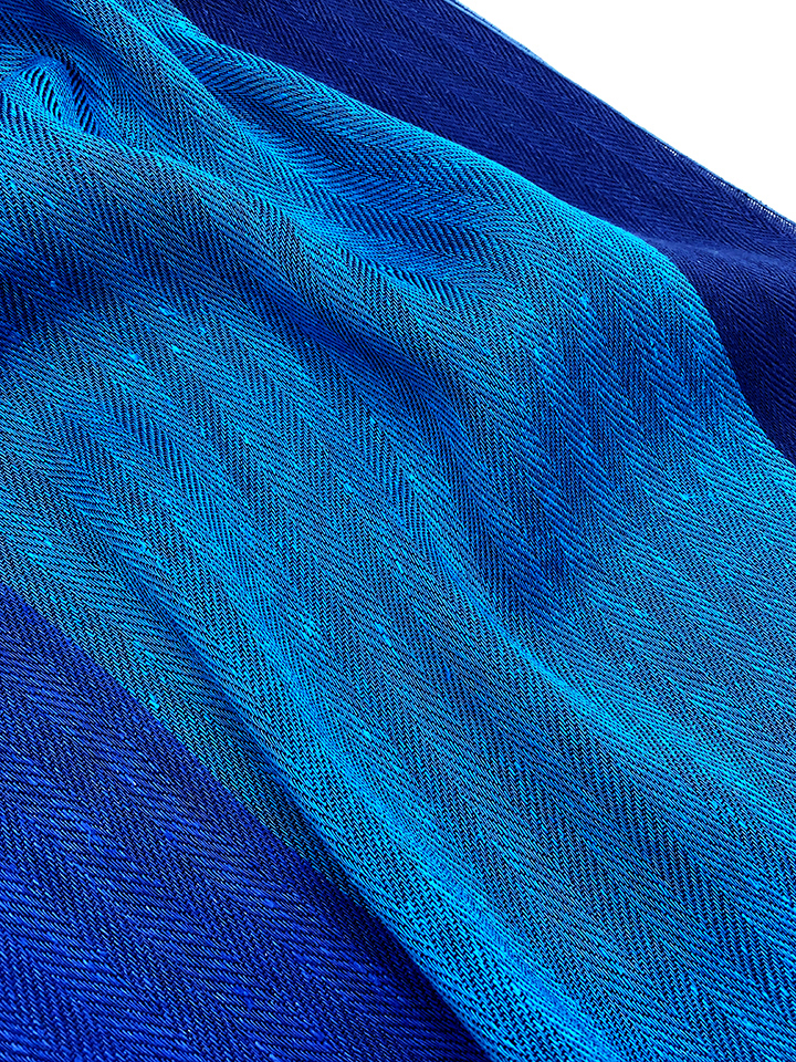 Ein breiter Schal aus blauem Leinen mit einem sanften Farbverlauf von dunkelblau zu hellblau und einem zarten Fischgrätenmuster.