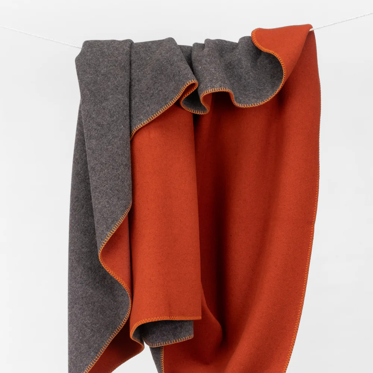 Die Merinodecke »FireSideMoon« von COASTLAND ist aus feiner, unbehandelter Merinowolle gefertigt und misst 150 x 220 cm. Sie zeigt eine Seite in Anthrazit und die andere in leuchtendem Glutrot. Die Decke ist dick, weich, dekorativ abgekettelt und dekorativ drapiert vor weißem Hintergrund.