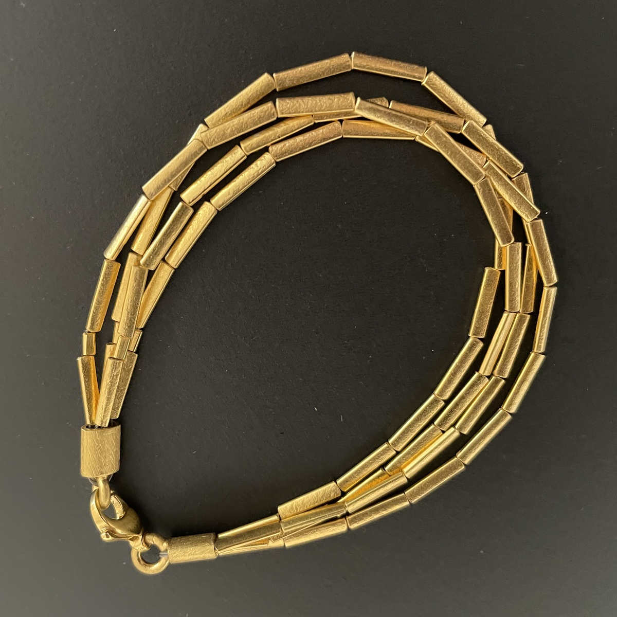 Dreireihiges Armband mit feinen, zylindrischen Gliedern aus Silber, feingoldplattiert, auf schwarzem Hintergrund, modernes und elegantes Schmuckstück für besondere Anlässe.