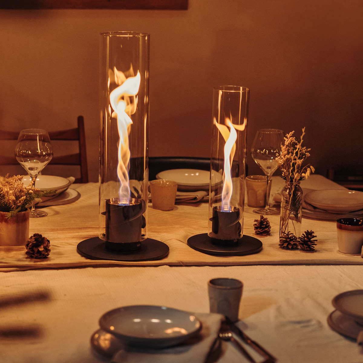 Gemütlich gedeckter Tisch mit Geschirr und zwei brennenden Tischfeuern (SPIN 900) unter Glaszylindern im Innenraum. Die warmen Lichtstrahlen der Flammen sorgen für eine stimmungsvolle Beleuchtung.