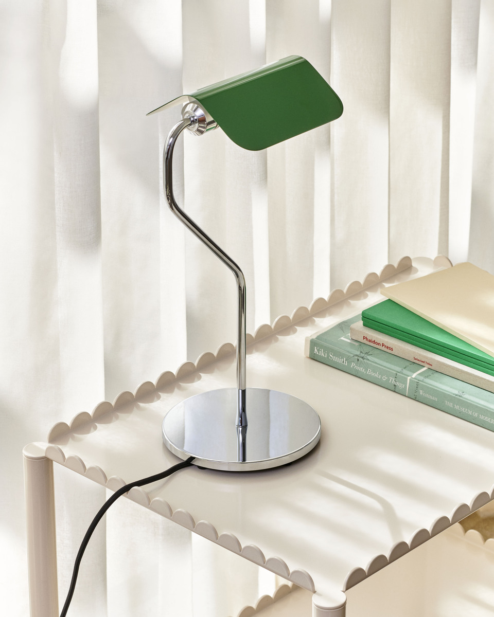 Eine moderne Tischlampe mit einem grünen Lampenschirm und einem eleganten gebogenen Edelstahlarm steht auf einem kleinen Tisch. Im Hintergrund sind geschlossene weiße Vorhänge zu sehen.