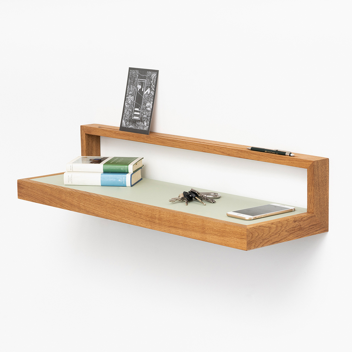 Wandregal mit Holzkorpus und pistaziengrüner Ablagefläche, geeignet für Bücher und Dekorationen, montiert an einer weißen Wand, mit darauf liegenden Alltagsgegenständen wie einem Buch, einer Postkarte und einem Smartphone.