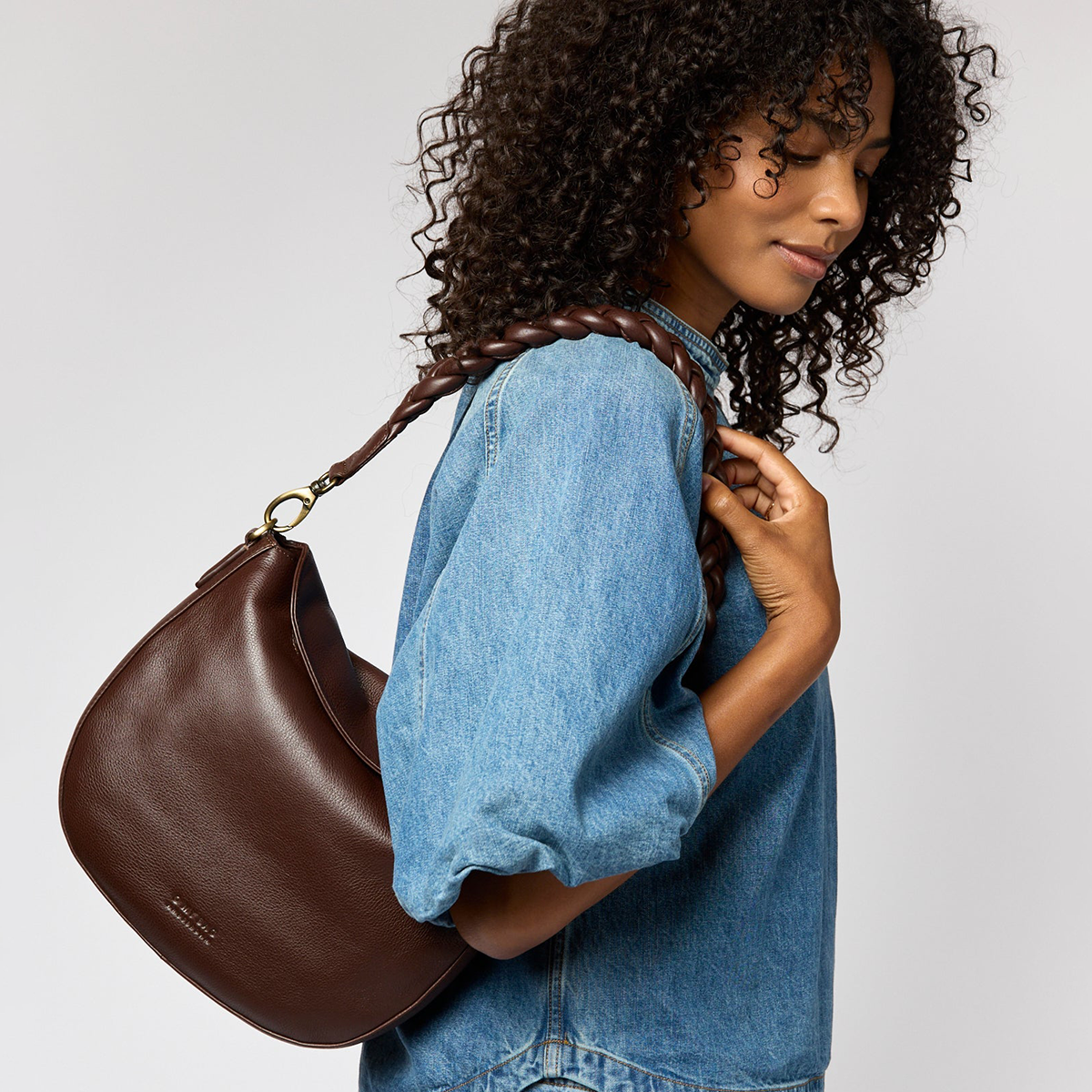 Frau trägt eine dunkelbraune Schultertasche aus Leder namens »Lana« über der Schulter. Die Tasche hat ein schlichtes, minimalistisches Design und wird zu einer blauen Jeansbluse kombiniert. Das Bild stellt die Tasche als modisches Accessoire für den Alltag dar.