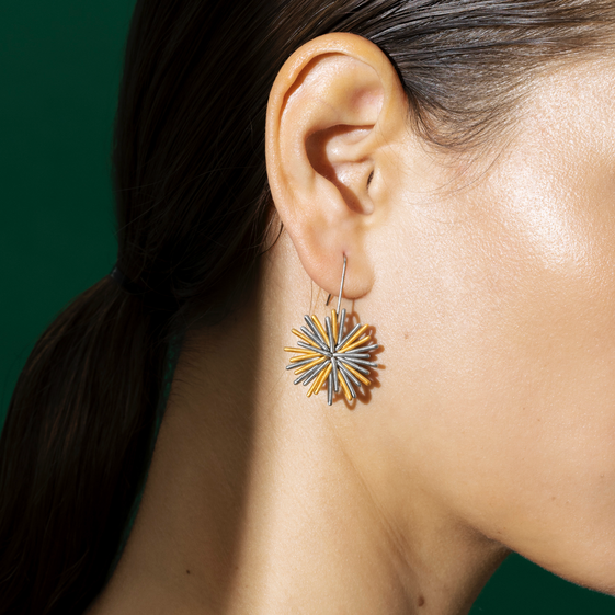 Detailaufnahme eines weiblichen Ohrs mit einem auffälligen, blumenförmigen Ohrring in Gold, Blau und Silber vor grünem Hintergrund. Das Schmuckstück wirkt modern und künstlerisch und ist für Damen geeignet.