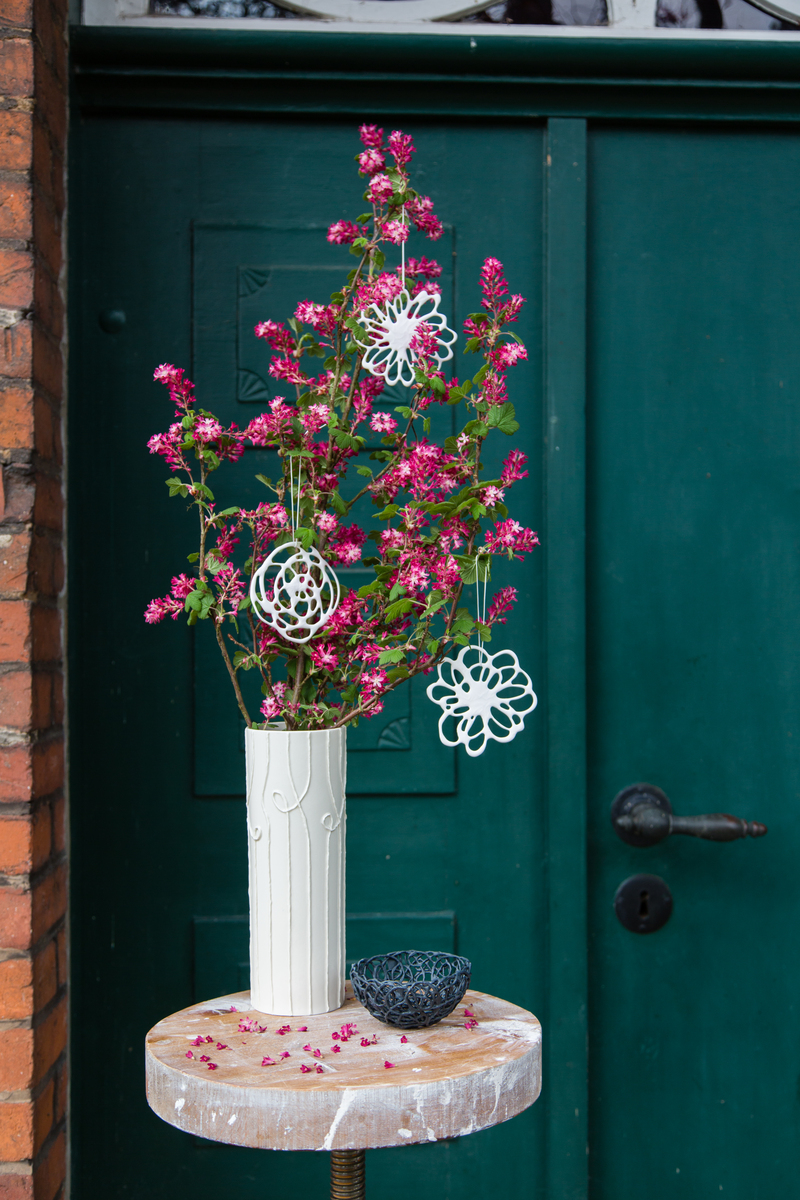 Ein sechsteiliges Set von handgemachten Porzellanblüten hängt an blühenden Zweigen in einer eleganten, weißen Vase. Die Blüten sind weiß und detailreich gearbeitet, während die Zweige lebendige rosa Blüten tragen. Die Szene ist vor einer grünen Tür platziert und zeigt eine Mischung aus Natur und handgefertigter Kunst.