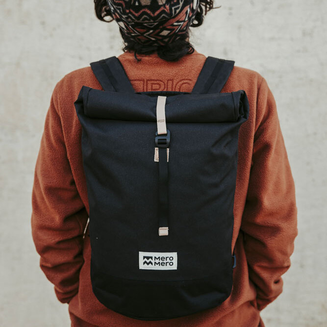 Person trägt eine schwarze Fahrradtasche/Rucksack »Annecy« auf dem Rücken, mit einer orangefarbenen Jacke und einer gemusterten Mütze.